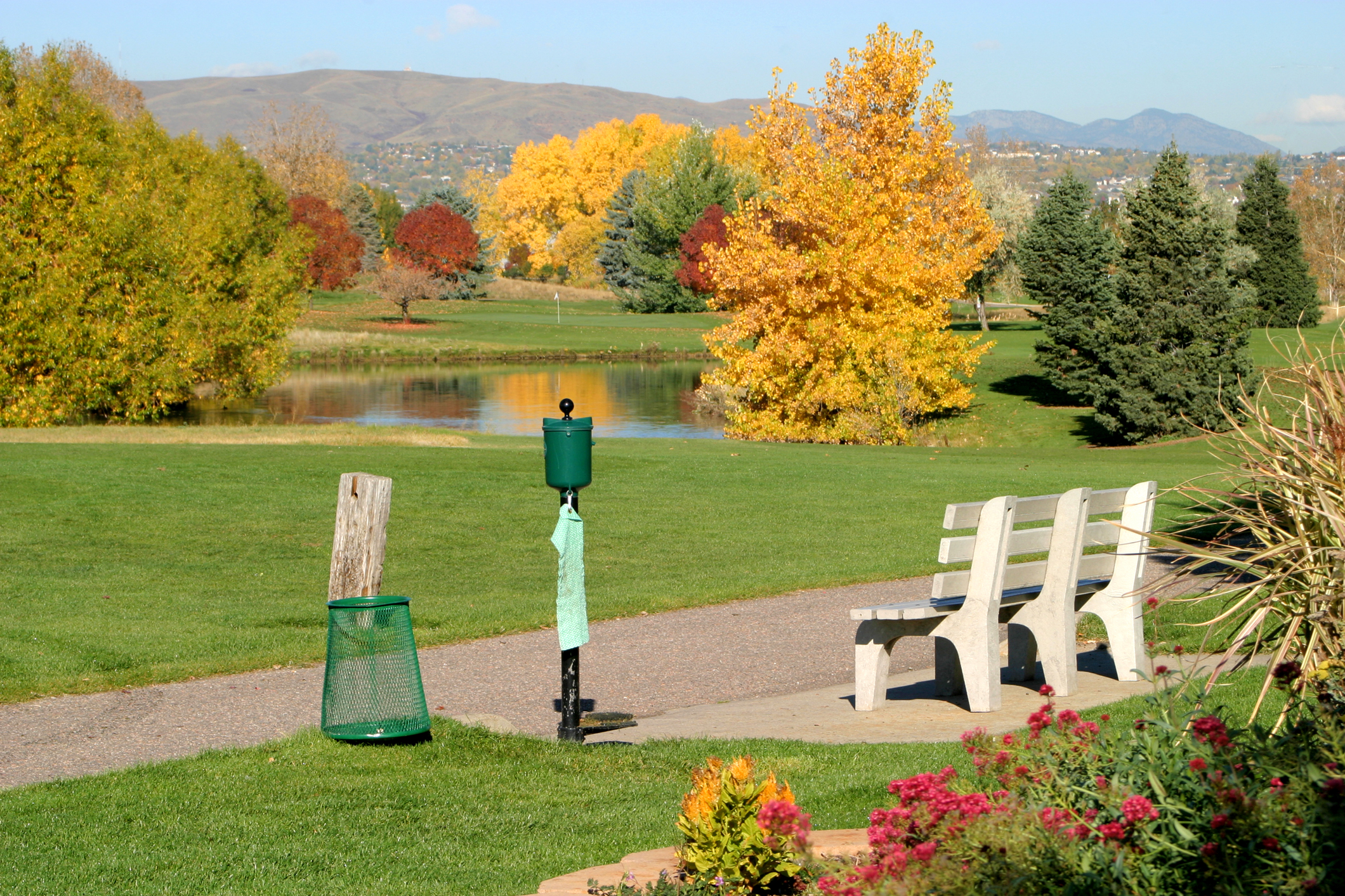 A bench, ball washer and trashcan near a tee box with fall foliage, trees and mountains in the background