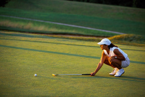 A woman crouched down on a golf green holding a putter assessing the line to putt her ball