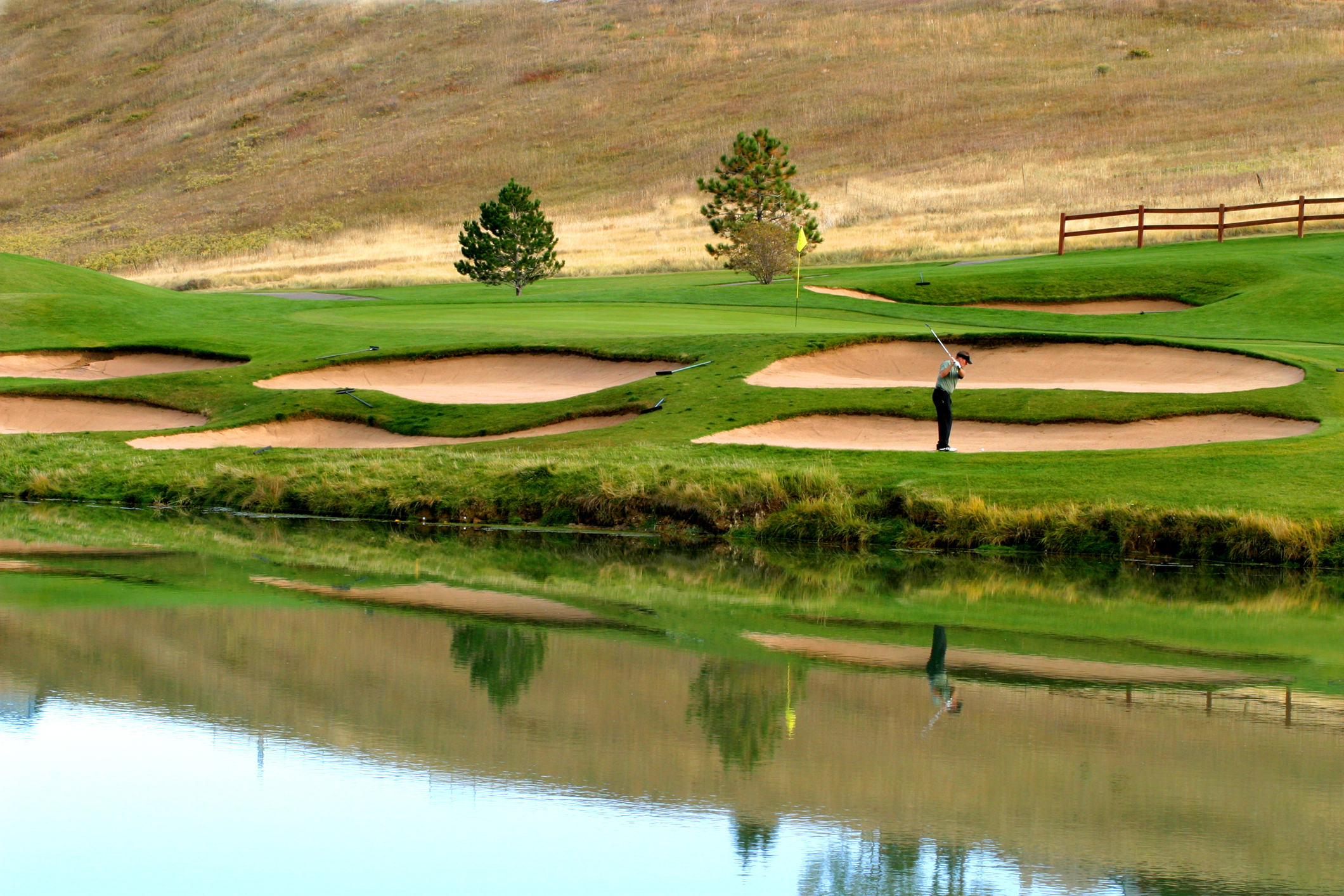 A golfer in mid-stroke getting ready to hit a ball in front of a golf green with water and bunker features