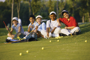 A group of smiling kids holding golf clubs sitting together on a practice green